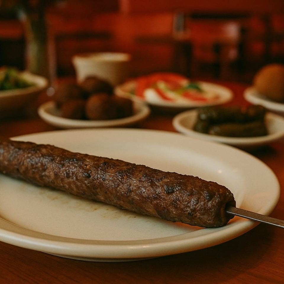 A grilled Mediterranean kofta kebab on a white plate, with various appetizers blurred in the background.