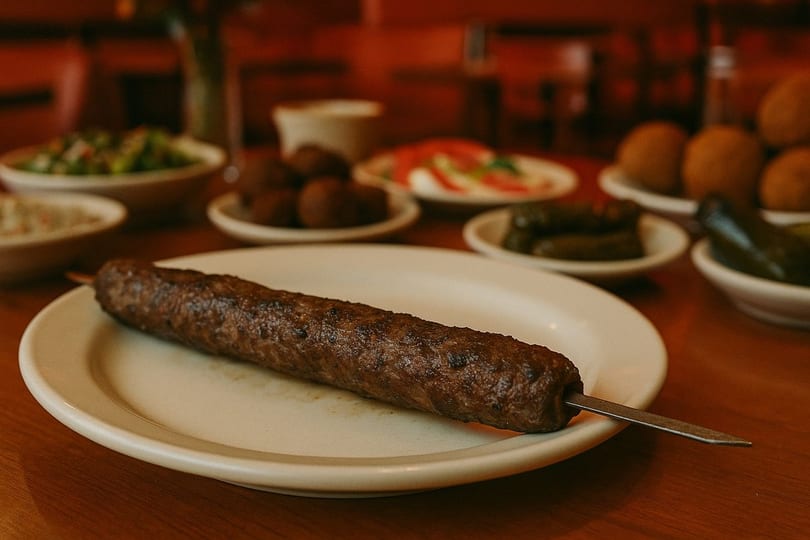 A grilled Mediterranean kofta kebab on a white plate, with various appetizers blurred in the background.